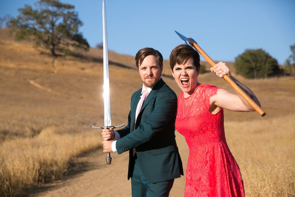 A man in a suit holding a sword stands next to a woman in a lacy dress who is brandishing an axe. They’re outside in a mostly brown field. 