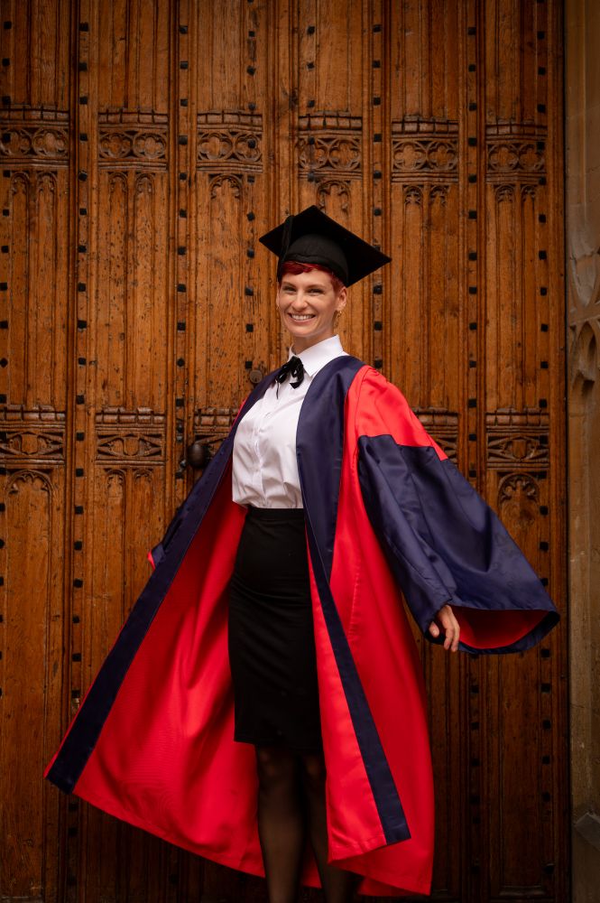 A woman in a red and blue DPhil gown twirling in front of a wooden door