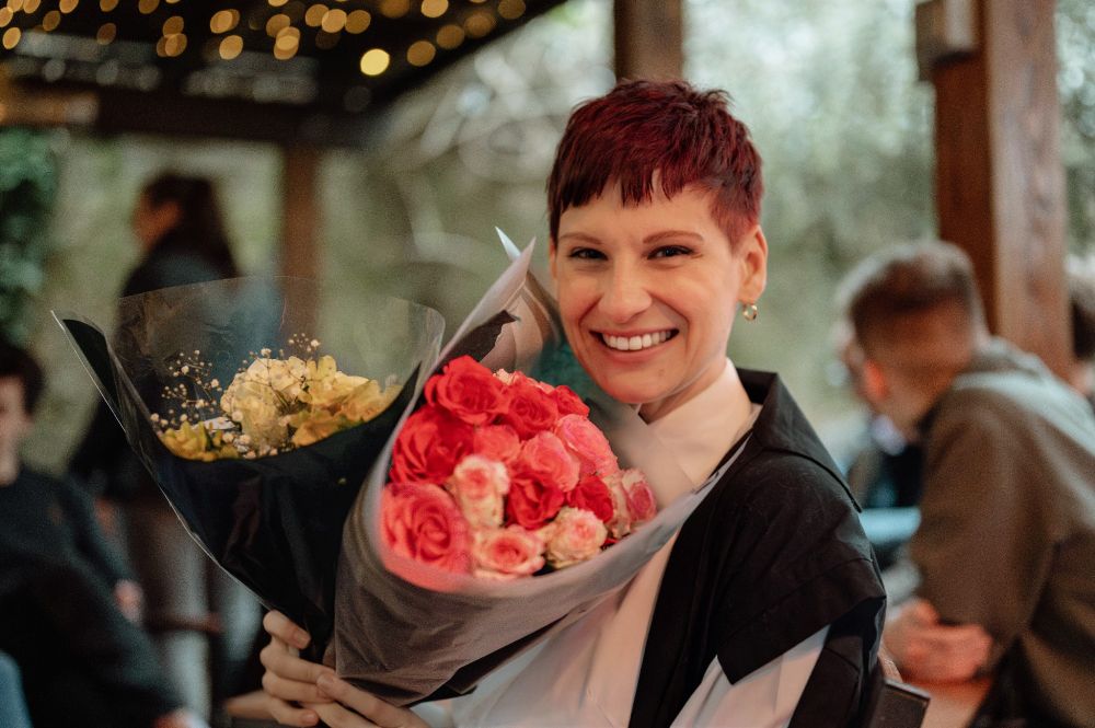 A woman in academic dress smiling whilst holding a bouquet.