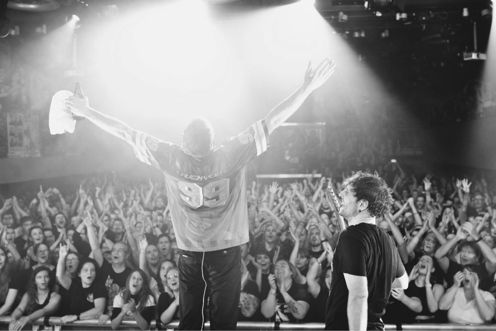 Konzertfoto der Band ITCHY. Im Vordergrund der Schlagzeuger der Band, der beide Arme ausstreckt. Daneben der Sänger und Gitarrist. Im Hintergrund das jubelnde Publikum.
