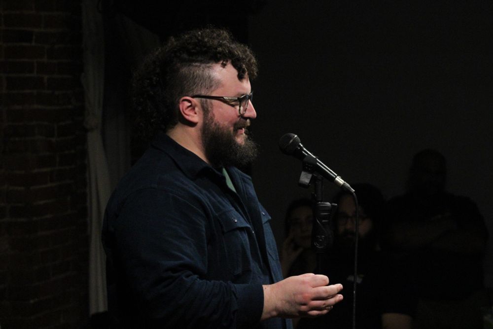 From the eyes down, a stereotype of a new Englander, in a blue LLBean thick flannel shirt, stands at a microphone during a poetry reading.  Atop his head is a curly mullet that has the shape of a horse's mane.