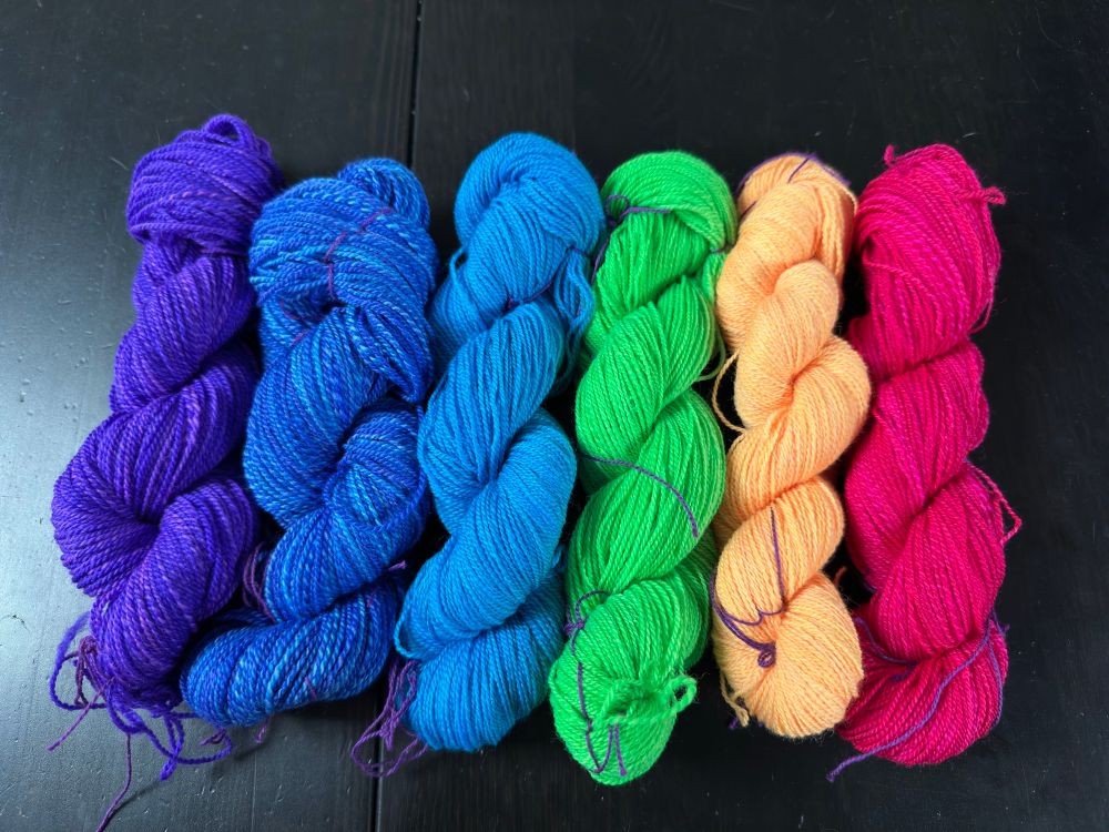 Six skeins of #handspun yarn in a rainbow. From left to right they are purple, dark blue, light blue, green, yellow, and pink. The skeins are much more cooperative than that bobbins to photograph. No tricks were employed to wrangle the yarns for this photo except minimal efforts to minimize the skein ties, which are purple cotton 2/8 weaving yarn. 