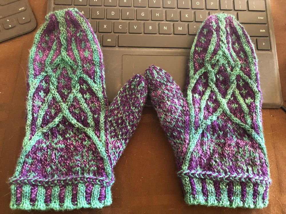 A pair of mittens sit on a computer keyboard. The mittens have a purple background with turquoise cables and colour work. The colourwork forms the shape of snowflakes in the background of the cables and there is a diamond pattern over the thumbs. The mittens are finished with two colour 2x2 ribbing. Pattern is Norrlund by Sarah Huntington Burch. 