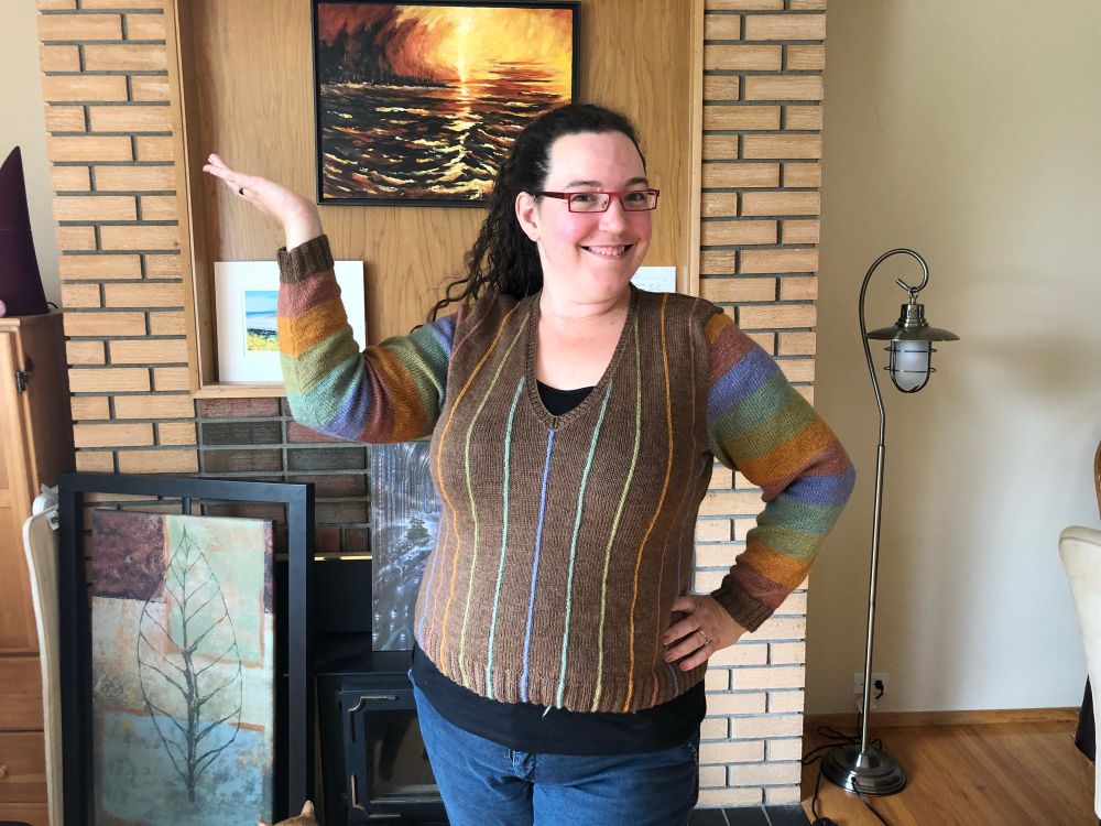A white woman (me) with dark hair and eyes and red glasses is standing in her living room showing off the sweater she just knit for herself. She has her left hand on her hip and is holding her right hand up in a “ta-da” kind of pose. The sweater is brown v-neck with narrow vertical stripes in earthy blue, green, orange, and pink. The sleeves have wide horizontal stripes in the same colours. In the background a brick fireplace, lamp, and leaf artwork are visible. 