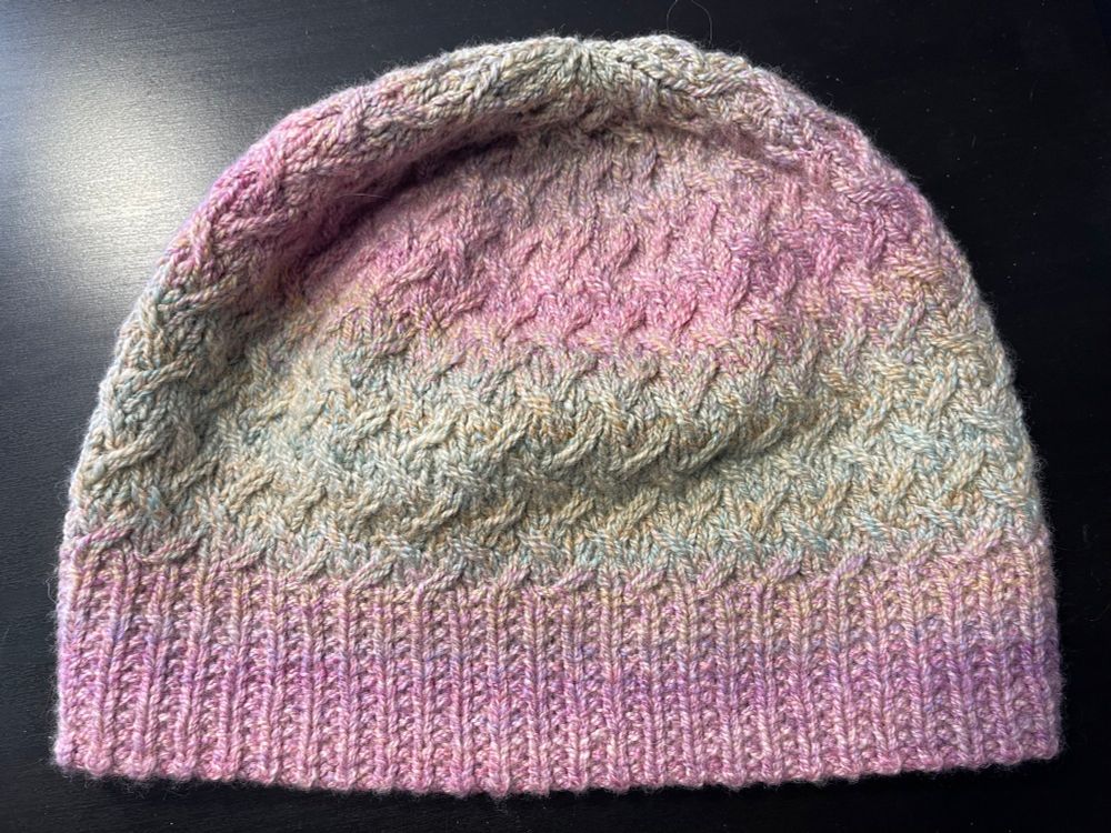 A toque made of handspun yarn that gently transitions from pink at the brim through a gentle yellow-green to pink again at the top. The hat has an allover cable pattern on it. The pattern is Field of Wildflowers by Joji Locatelli. The yarn is a 4-ply cabled yarn. 