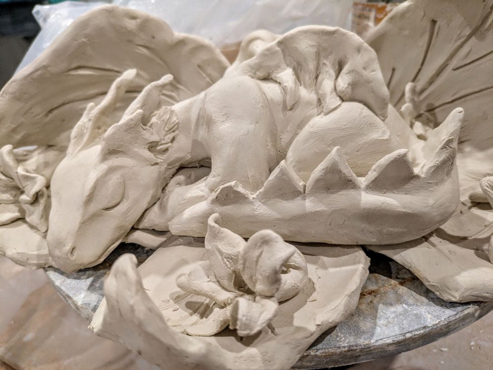 A sleeping baby dragon on a nest of leaves with dropped flowers around him, all sculpted out of English porcelain 