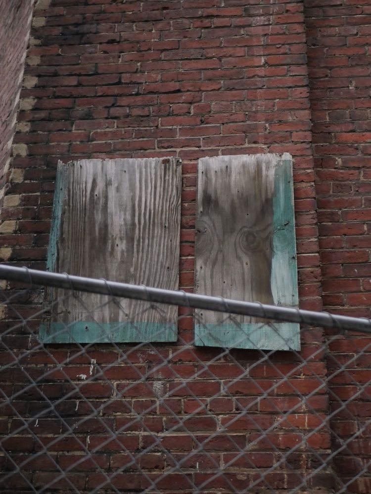 wooden boards attached to the outside of an abandoned brick building, through a chain link fence. it appears that a sign was at some point attached to them - there is a paint outline - but what, who knows.