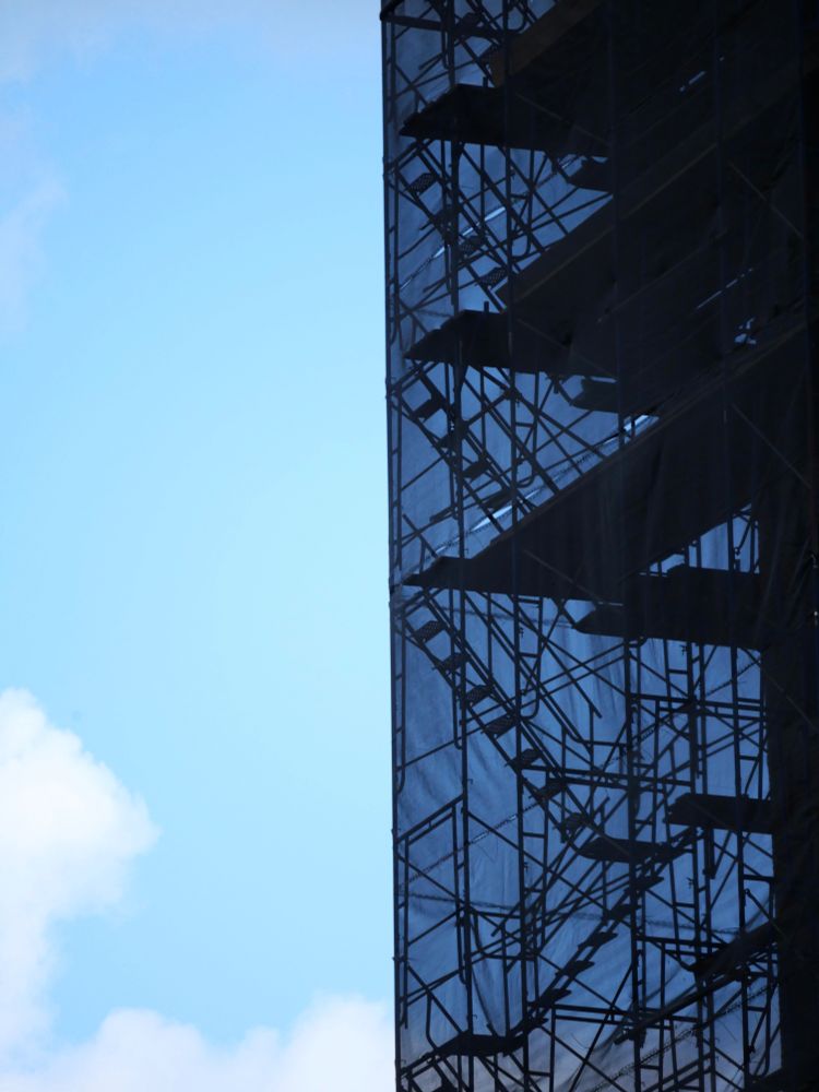 a scaffolded tower wrapped in semitranslucent fabric darkening the sky blue behind it, set against a much brighter sky