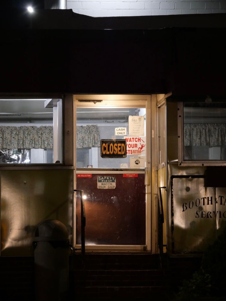 A closed sign, lit from above, on the door of a diner at night. The more you look, the more you think this places is, like, CLOSED-closed