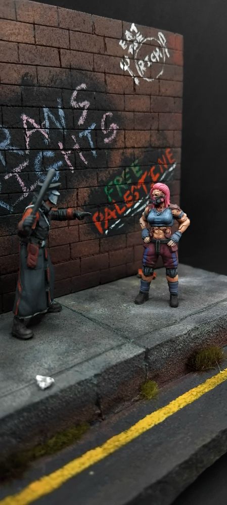 A woman standing off against two futuristic police all standing on a pavement with rubbish on it and a wall which has assorted graffiti saying "trans rights" "free Palestine" "eat the rich" "fuck terf island" "acab" and "z <3s a" 