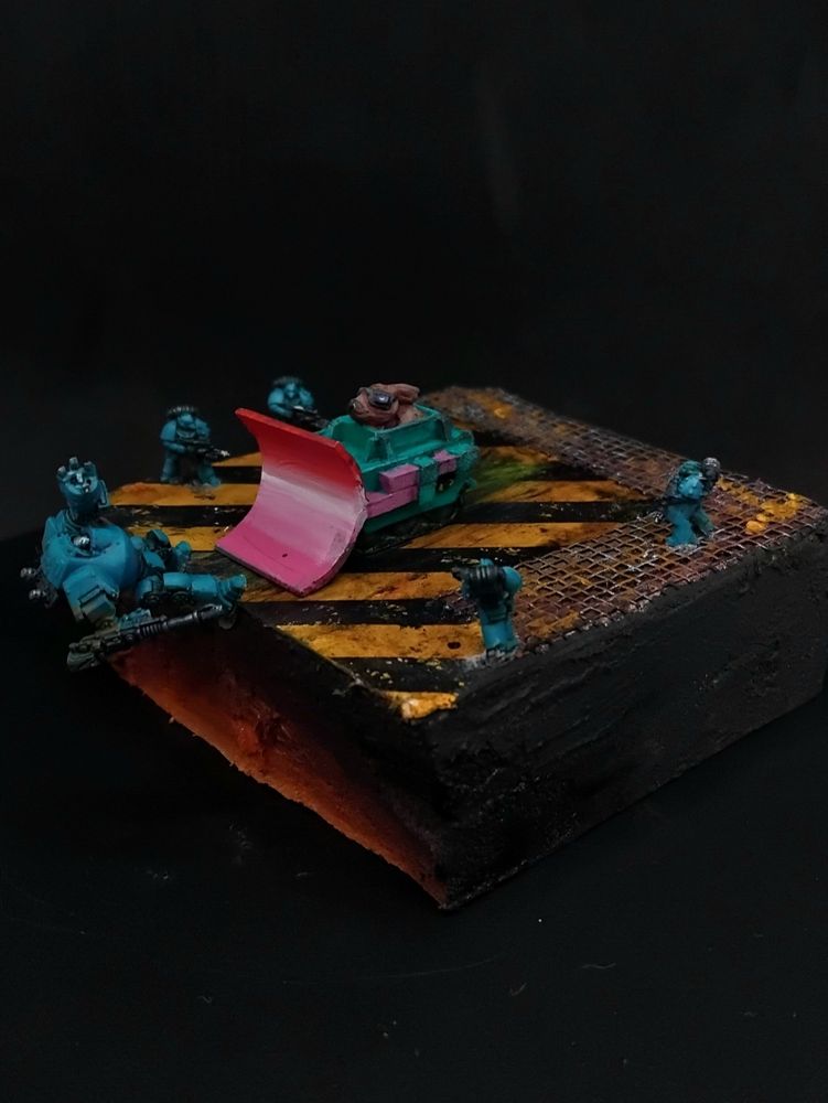 Rabbit in a vehicle with a bulldozer front (painted like the lesbian flag) pushing a dreadnought off the side of the diorama into something hot as marines close in