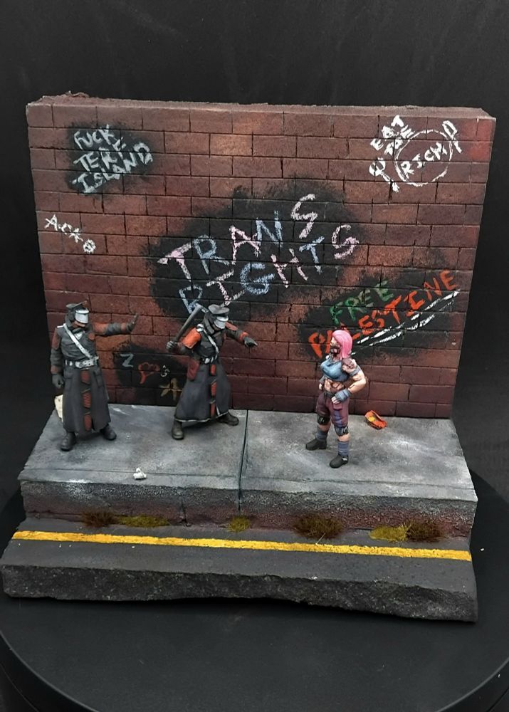 A woman standing off against two futuristic police all standing on a pavement with rubbish on it and a wall which has assorted graffiti saying "trans rights" "free Palestine" "eat the rich" "fuck terf island" "acab" and "z <3s a" 