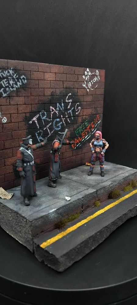 A woman standing off against two futuristic police all standing on a pavement with rubbish on it and a wall which has assorted graffiti saying "trans rights" "free Palestine" "eat the rich" "fuck terf island" "acab" and "z <3s a" 