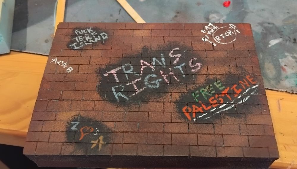 A foam block cut to look like bricks painted as redbrick with graffiti on it saying "fuck terf island" "free Palestine" "acab" "trans rights" "eat the rich" and " z <3 a"