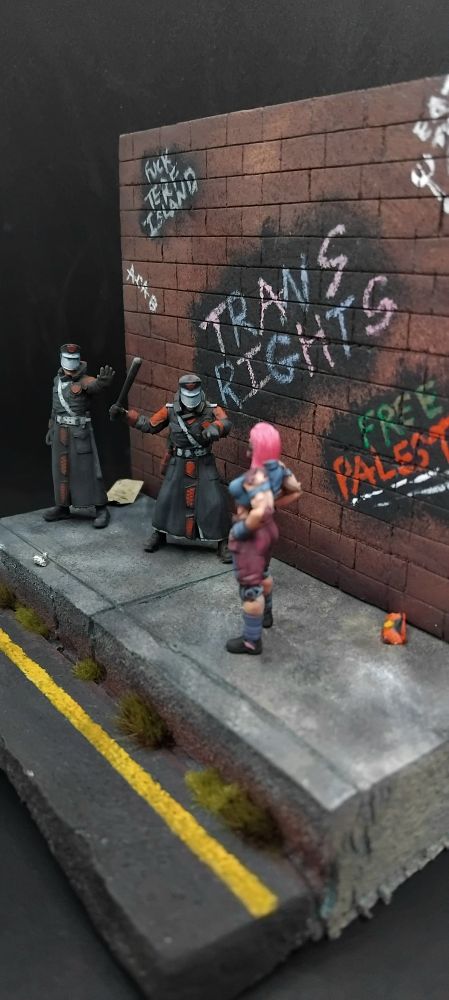 A woman standing off against two futuristic police all standing on a pavement with rubbish on it and a wall which has assorted graffiti saying "trans rights" "free Palestine" "eat the rich" "fuck terf island" "acab" and "z <3s a" 