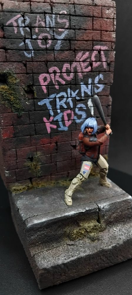 Person with blue hair, cyber legs, red jacket and jeans wielding a baseball bat infront of a wall that say 
