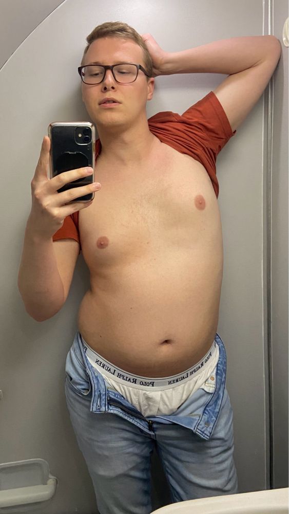Me in an airplane restroom showing off my naked torso. My shirt is tucked behind my neck, my left hand is behind my head, and my pants are unzipped showing off my boxers.