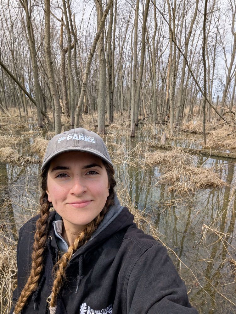 a selfie of me in winter gear, standing in a forested wetland. There are trees and old reed canary stems in the background. 