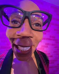 selfie com zoom cómico no rosto do rupaul, está usando um óculo quadrado preto com armaduras grossas, um blazer xadrez e ao fundo uma parede de tijolinhos rosas