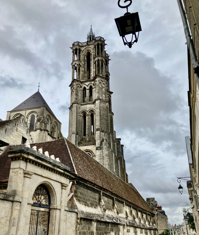 Top of image L-R romanesque tower topped by grey tiles. Next an ornate early French gothic tower with 4 layers visible above the red-tiled roof of the cloister. This tower is topped by a small campanile. Foregrounding the top right of the image is street lamp hanging from a curled, iron wall holder. Bottom left is a gateway into the cloister containing a black, iron double gate. The buildings are both made of light coloured stone, darkened by age and pollution in places