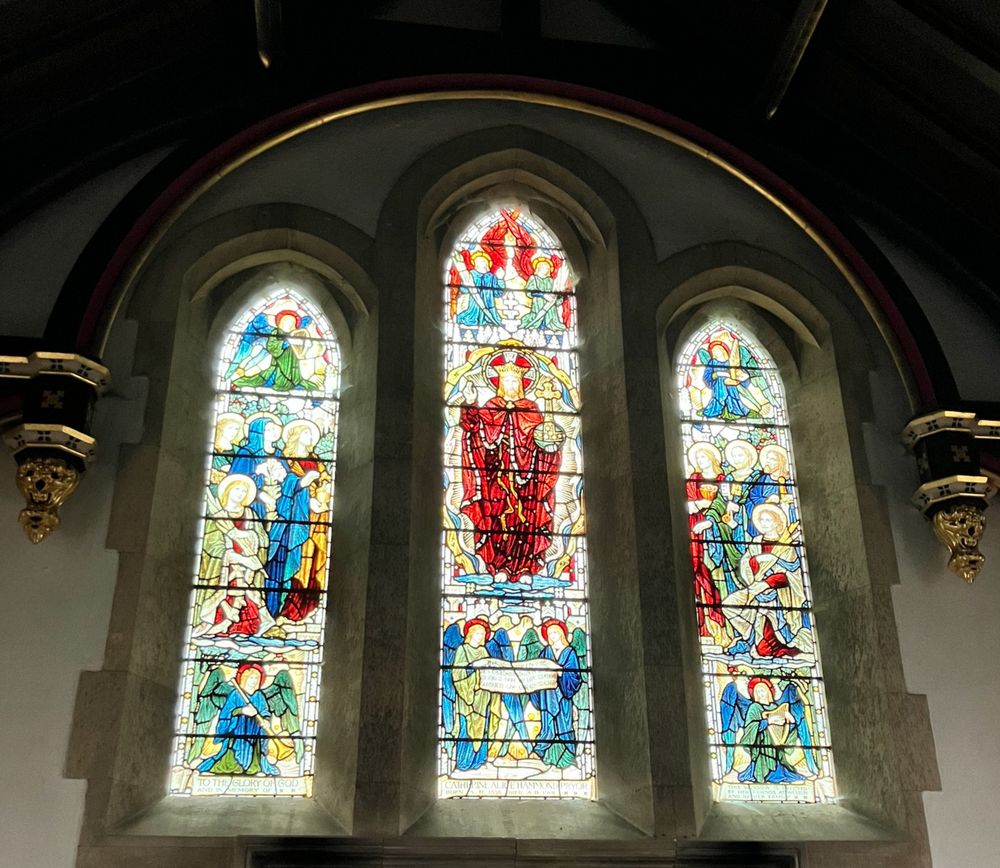 Three light, stained glass, east window of the church. The images are of the Adoration of Christ Ascended. The central light shows Christ in red robes, crowned and holding an orb. The glow-up comes from the way the light is hitting the glass, giving a haloed effect.
