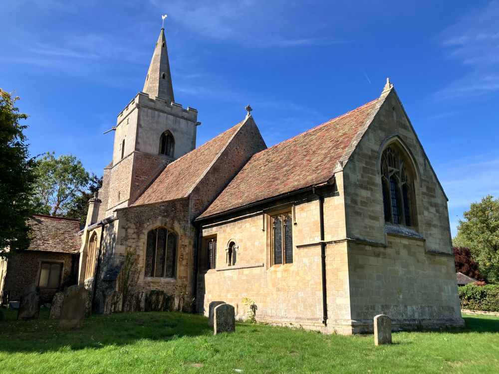 Medieval church, renovated by the Victorians. Photo taken standing in the churchyard shows from right to left chancel with east end window, nave, south aisle with large late medieval window, tower with top 2 stages visible. Top stage has crenellations, with a steeple topped by a shiny, cockerel weather vane. Bright blue last-of-summer sky and a warm 27 degrees.