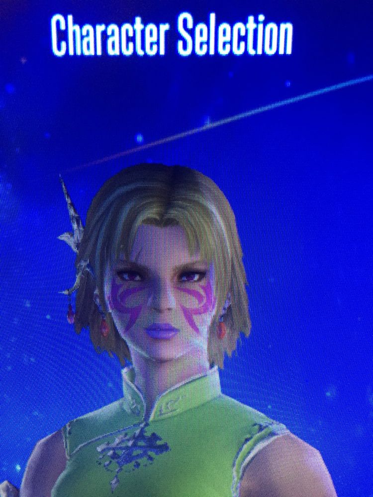 A blonde woman with pink facial markings, wearing a green dress in front of a blue background.