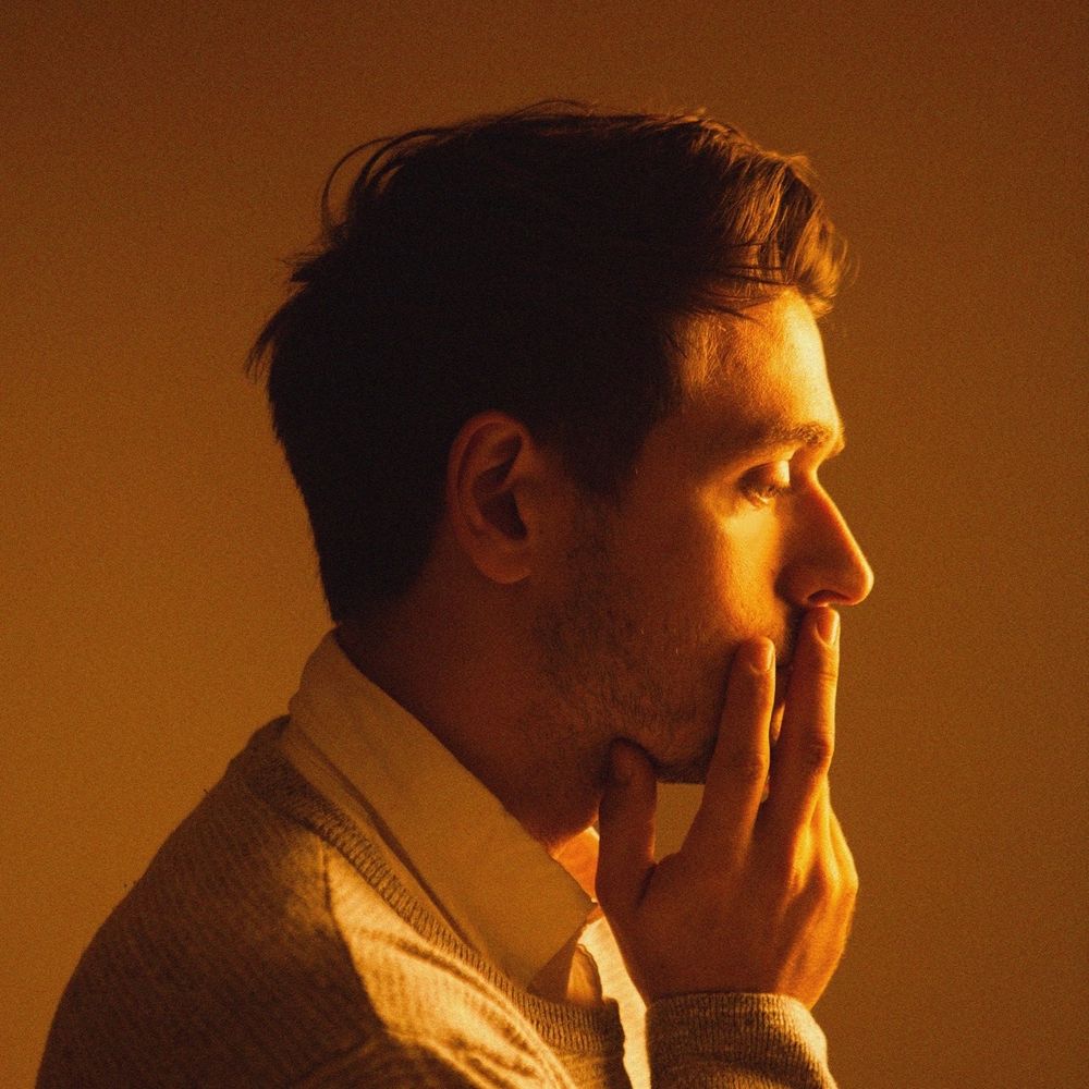 An orange-lit young white man with short hair in profile, with his hand on his mouth