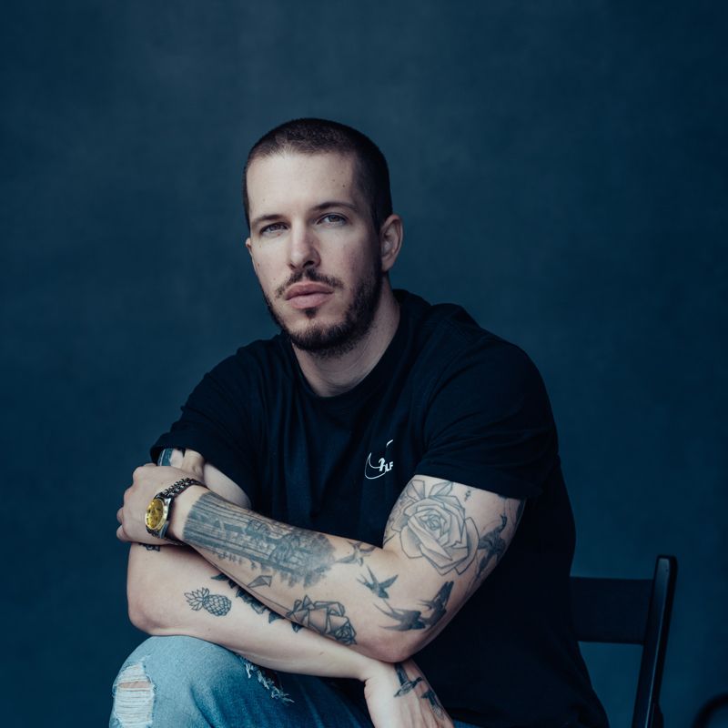 A young white man with close-cropped hair, beard, and mustache with arm tattoos and a gold watch wearing a short-sleeved black shirt and sitting in a chair