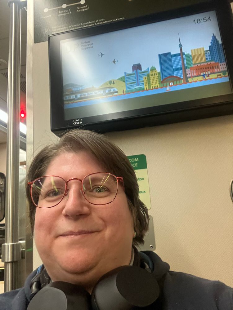 Me in the UP Express, the train going to Toronto city centre from the airport. Behind me is a screen showing a cartoon version of Toronto skyline.