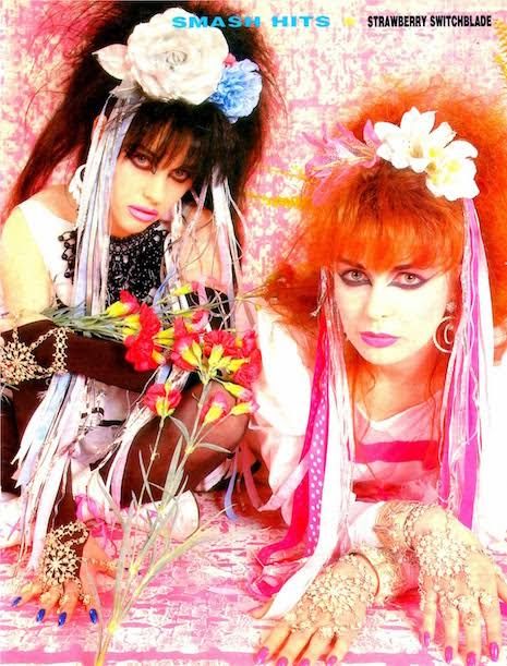 2 women, 20s, a redhead and a brunette in close up. They're leaning on a pink table in front of a pink backdrop. They are wearing headpieces with roses and ribbons,their hair is extravagantly teased.