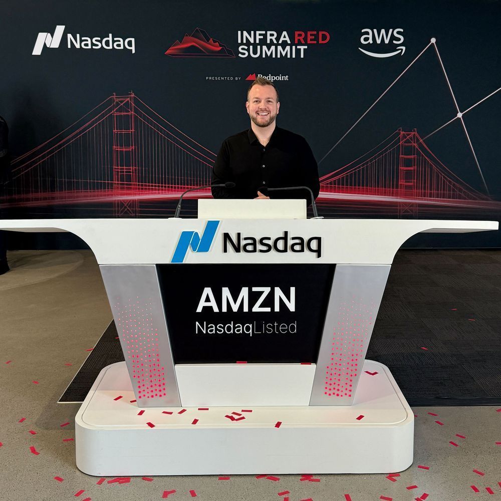 Rick stands smiling at the Nasdaq closing bell podium with AMZN (Nasdaq Listed) displayed on its screen. Behind him is a backdrop featuring the Golden Gate Bridge in red outline, along with the logos of Nasdaq, InfraRed Summit (presented by Redpoint), and AWS. Red confetti is scattered on the podium's base.