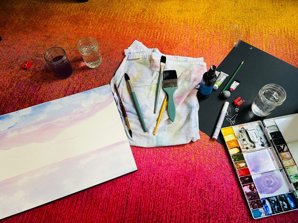 A watercolor block with light washes of blues and pinks on the far left side, a rag with paintbrushes in various sizes, and a palette of watercolors on the right. Cups of water just above the watercolor block, on top of a pink and orange rug. 