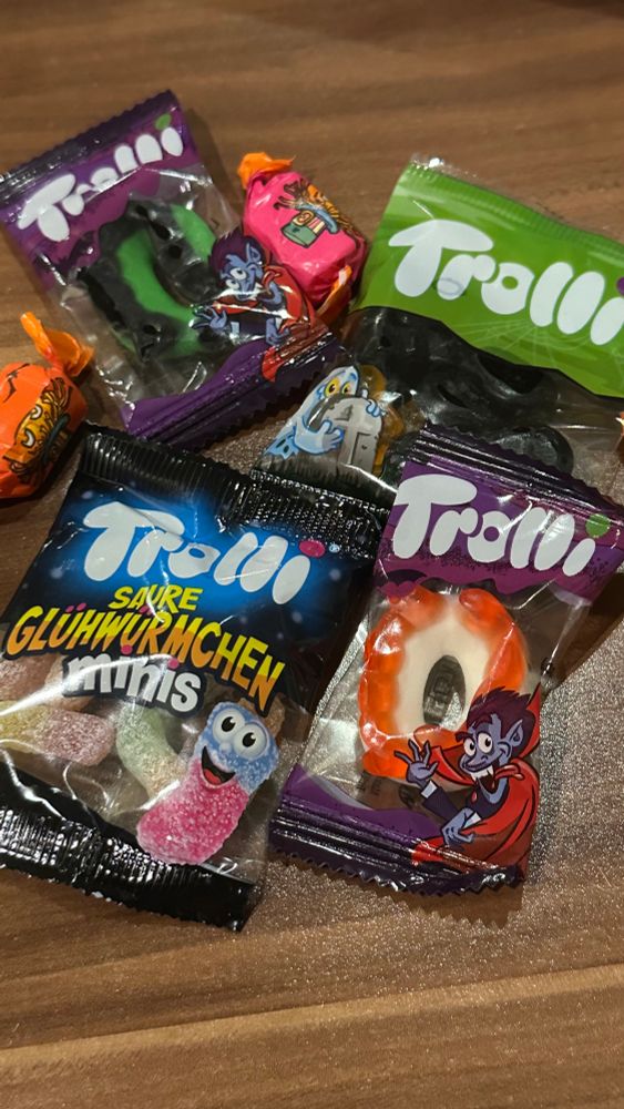 Auf einem Holztisch liegen Mini Tüten mit Gummi Süßigkeiten und Kaubonbons. Auf den Tüten steht Trolli, vorne liegen „saure Glühwürmchen Minis“, daneben und oben liegt ein Gummi Draculq Gebiss, oben rechts schwarze Lakritz. Auf den Kaubonbons sind Kürbisse abgebildet. 