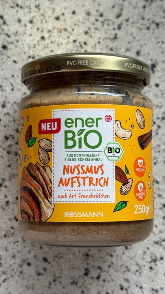 Ein Glas enerbio Nussmus Aufstrich nach Art Franzbrötchen. 
Das Etikett ist gelb, darauf abgebildet sind gezeichnete Zimtstangen, Mandeln, Cashews und Franzbrötchen. 