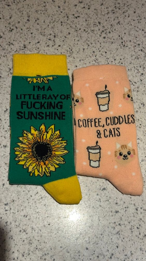 Auf einer gesprenkelten Oberfläche liegen zwei Paar Socken. Das linke Paar ist grün, hat gelbe Akzente und eine Sonnenblume darauf, darüber steht “I'm a little ray of fucking sunshine”. Das rechte Paar ist pfirsichfarben und hat Kaffee to-go Becher und Katzen abgebildet, mittendrin steht „coffee, cuddles & cats“. 