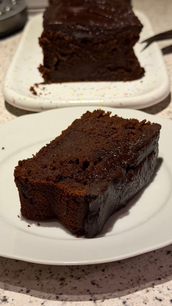 Auf einem weißen Teller liegt ein dunkelbraunes, rechteckiges Stück Schokokuchen. Dahinter ist auf einem bunt gesprenkelten Teller der restliche, lange Kuchen. Rechts vom Stück Kuchen glänzt die Fläche mehr. 