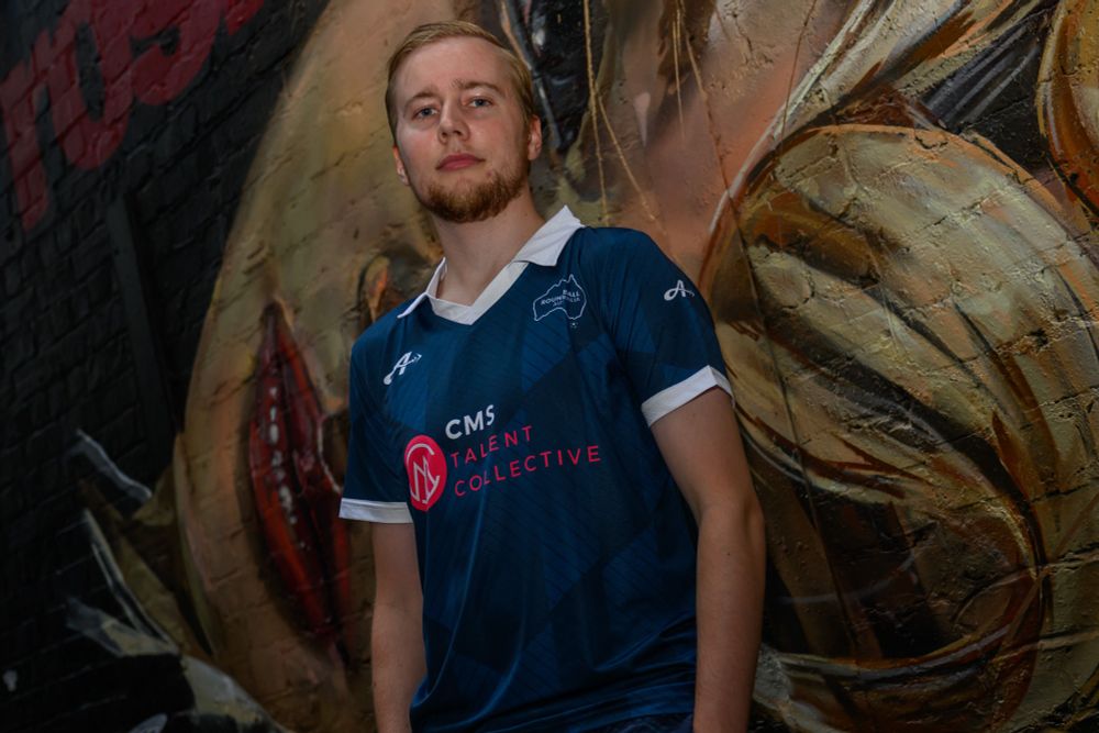 A man with short blonde hair wearing a blue jersey with a white collar and trim standing in front of a wall with mural artwork