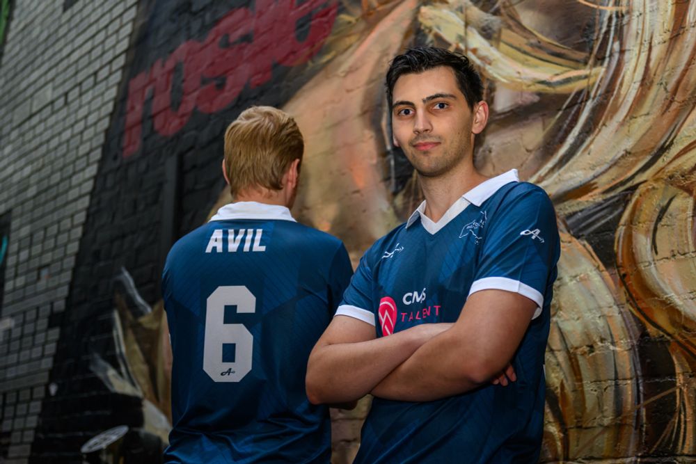 A man with black hair wearing a blue jersey with a white collar and trim standing in front of a wall with mural artwork, standing to the right of a man with blonde hair wearing a blue jersey with a white collar, staring towards a wall with mural artwork, with the jersey reading "AVIL 6" on the side facing the camera.