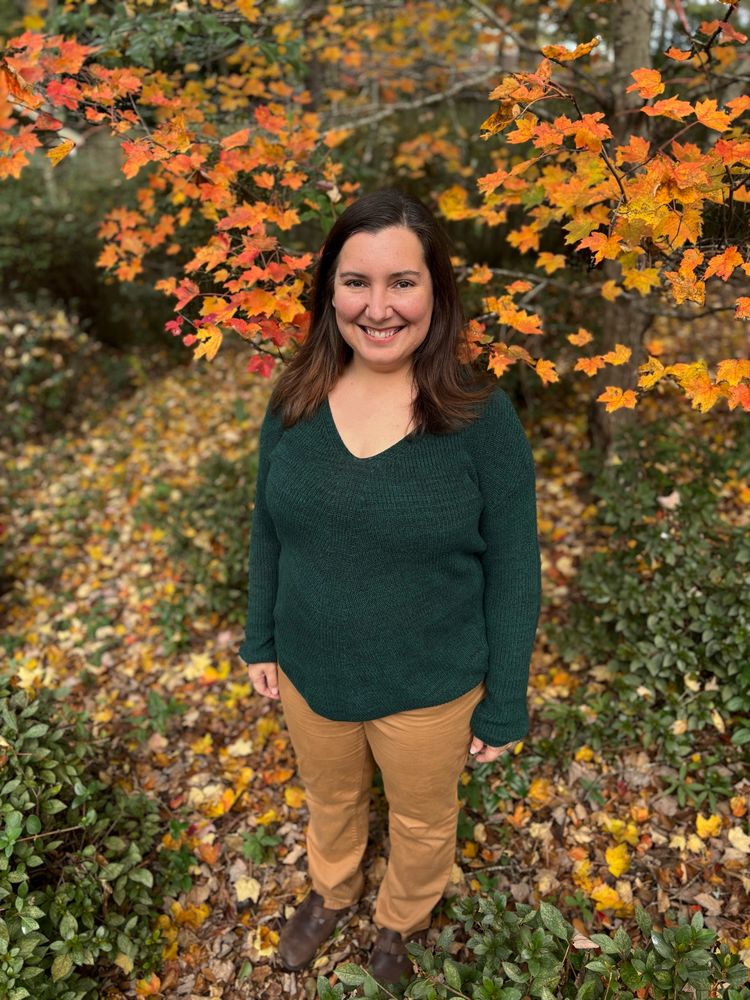 A woman with brown hair and a green sweater stands in front of a tree with fall colors. She is not sucking in her stomach but she is smiling brightly. 