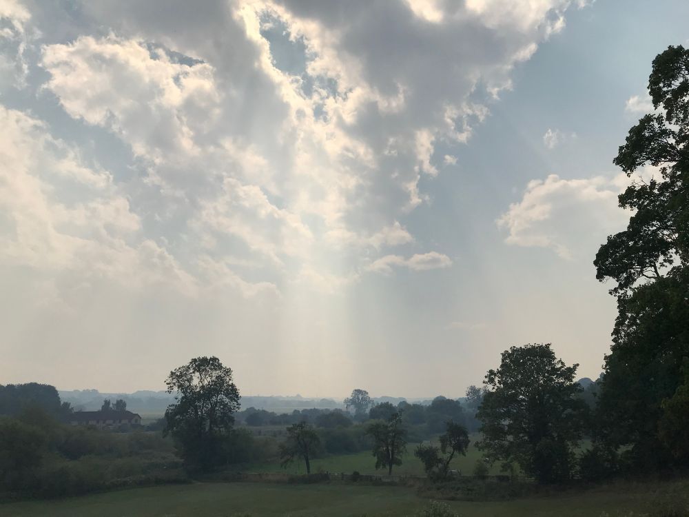 Rays of sunlight pierce through puffy grey clouds and smoke. A landscape of trees and fields made hazy by smoke from burning moorlands in the distance. 