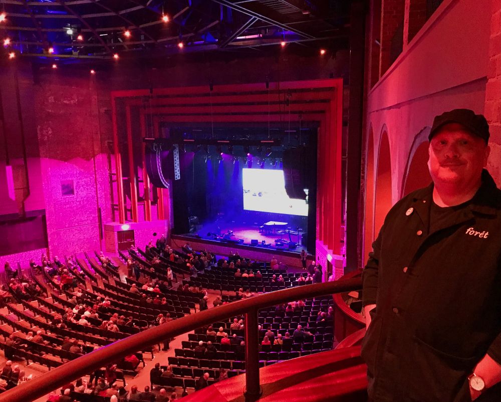 Me standing next to a railing overlooking an auditorium full of seating, a few patrons taking their seats. The room is lit in reds and pinks, the stage is blue with a brightly illuminated screen at the rear. I look like a French baker who enjoys his own bread a little too much (put the baguette down tubby). 