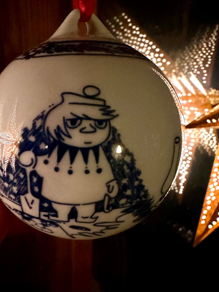 två senapsgula julstjärnor reflekteras i fönster och upphängd julgranskula i keramik, vit blå. där tootiki syns med en spade framför nån form av hög.