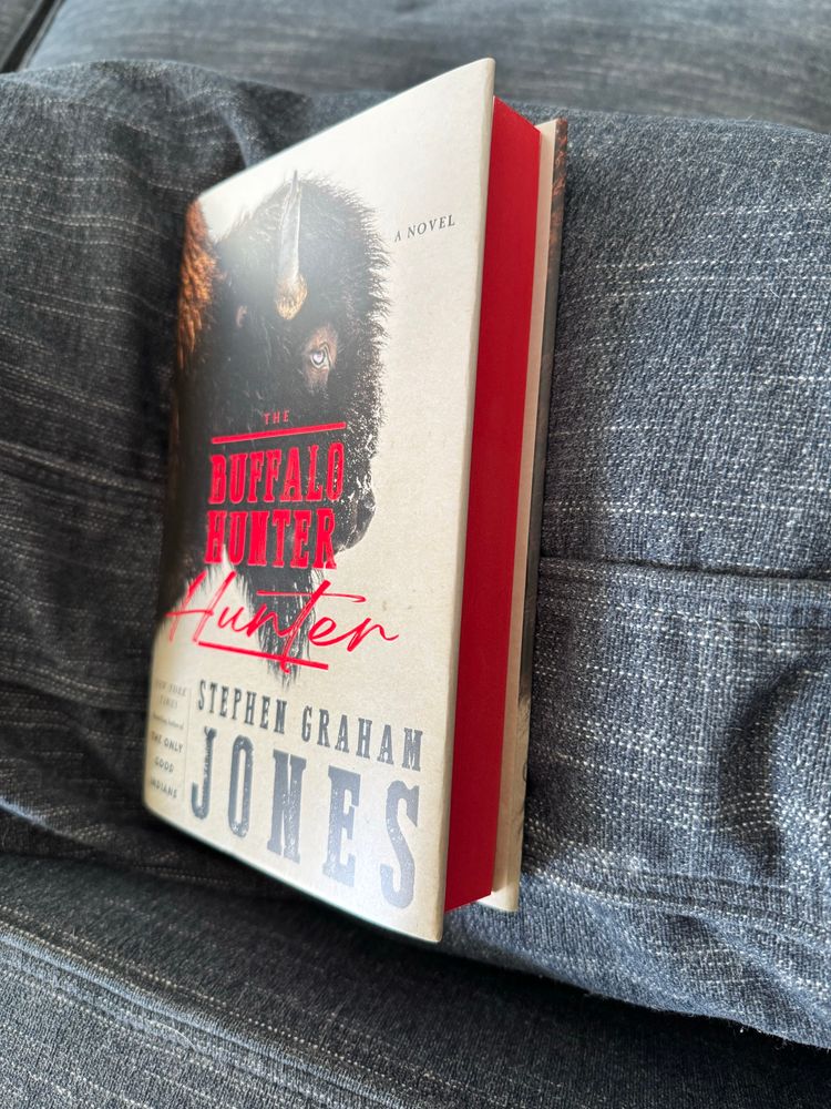 A copy of Stephen Graham Jones’s latest novel with red page edges resting on a blue couch cushion. 