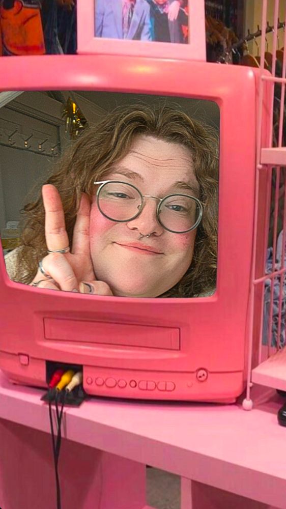 Me, Jules, a white transfemme with glasses, curly brown hair and rings on, framed in a pink CRT TV. I'm flashing the peace sign 