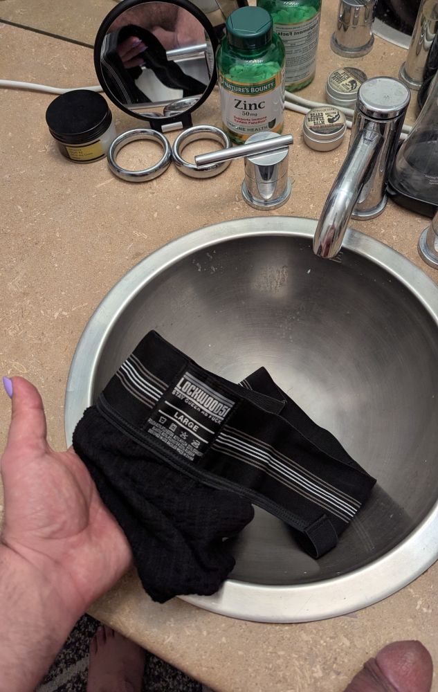 Photo of a bathroom sink and counter showing off a sweat soaked black athletic jock. In the bottom right corner of the frame is a half hard fat cock which is also visible in a countertop hand mirror next to a bottle of zinc supplements, steel cockrings, and facial moisturizer.