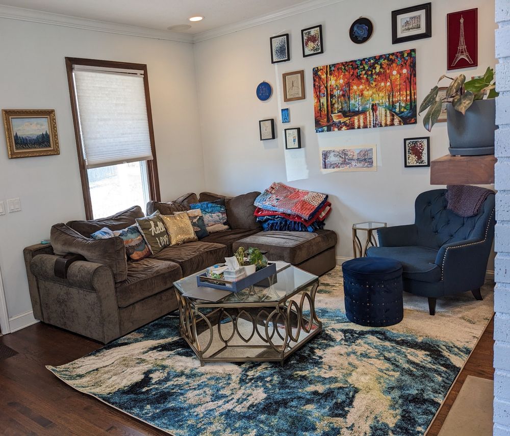 living room! this space has a beautiful area rug with blue, green, and beige swirls over hardwood, a large squashy gray sectional couch with lots of colorful throw pillows and blankets, a blue padded armchair with matching ottoman, a brass and glass hexagonal coffee table (and matching end table), and a gallery wall with lots of colorful art. walls are off-white and a nice big window is visible, partially obscured with a blind. there are two plants in this pic, one sad one in the middle of the coffee table, and one considerably happier pink princess philodendron in the foreground, on the mantle of the fireplace