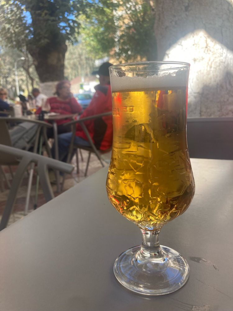 A glass of golden lager on a grey plastic table, there is a tree lined Spanish street in the background 