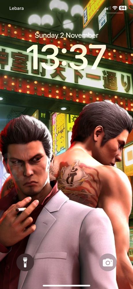 An iPhone XR lock screen, the art displayed on it depicting the original protagonist of the Yakuza video games, Kazuma Kiryu. He wears a light grey blazer and a dark red button-up collared shirt, and he’s holding a cigarette between his index and middle finger. Behind him is, well, him again but from the back and without his blazer and shirt on, with only some the top of his Yakuza tattoo visible due to being obscured by the first Kiryu. The background is the fictional city of Kamurocho.