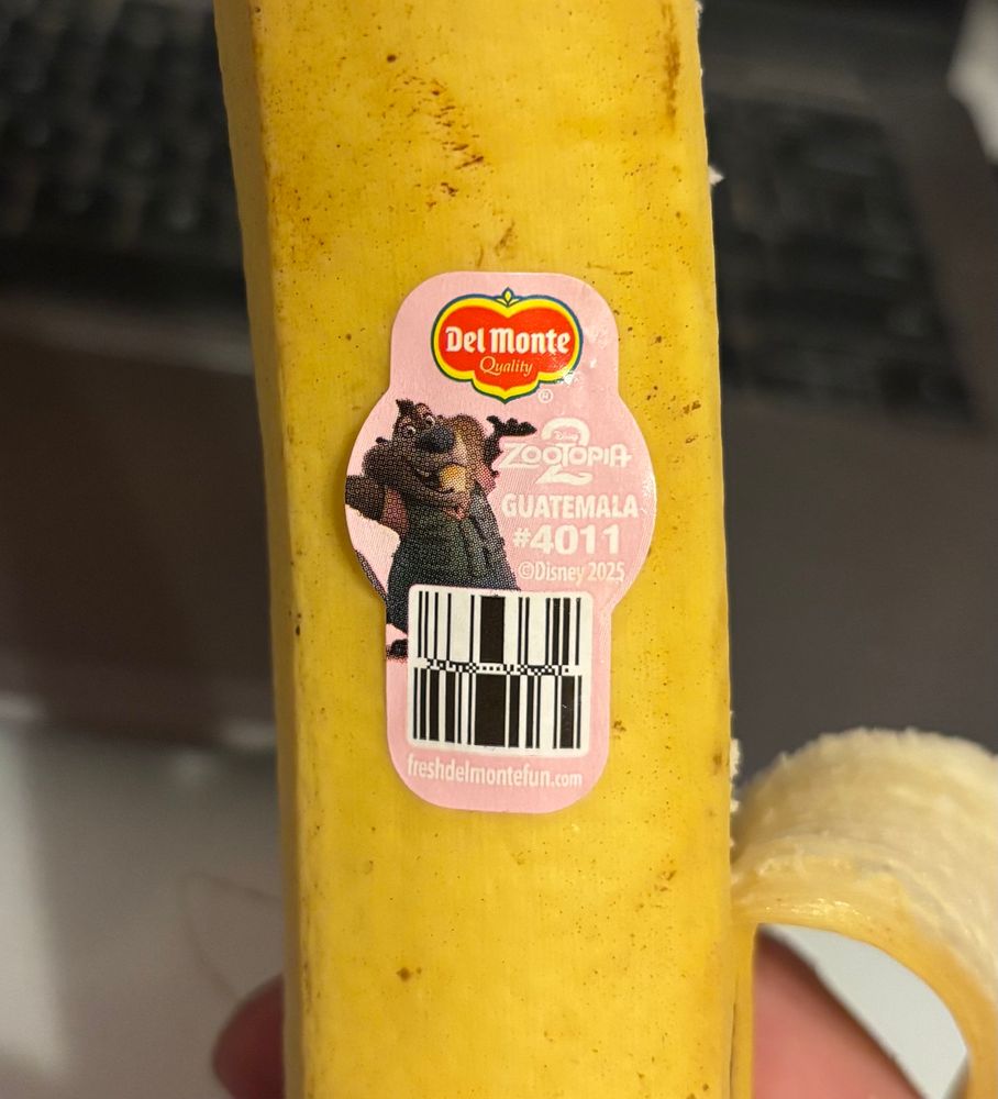 A close up on a banana bearing a sticker indicating that it was distributed by Del Monte and is a product of Guatemala. The sticker is pink and has an image of a cgi cartoon beaver on it. The beaver is Nibbles Maplestick, a character introduced in Zootopia 2.